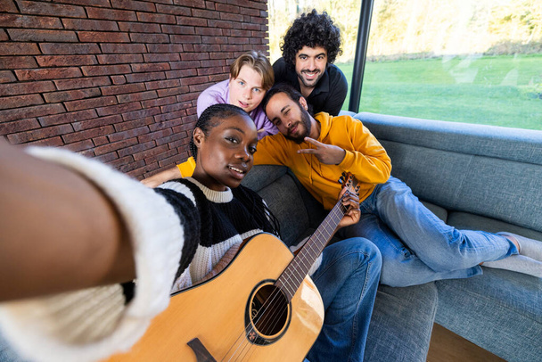 Ez az élénk kép négy különböző barát örömteli összejövetelét örökíti meg egy modern nappaliban. Egy fekete nő gitárral a kezében szelfit készít a barátaival, egy fehér nővel, egy közel-keleti férfival. - Fotó, kép