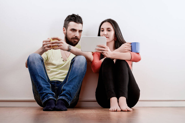 Счастливая пара на полу с кофейными кружками, радостно занимающаяся планшетом, их смех наполняет комнату - Фото, изображение