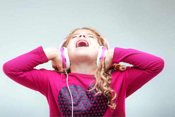Зухвала молода дівчина з кучерявим світлим волоссям, одягнена в рожевий колір, використовує білі навушники, пристрасно співаючи з блискучим виразом - Фото, зображення