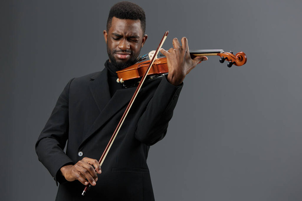 Ein talentierter afroamerikanischer Mann im schwarzen Anzug spielt Geige auf einem glatten grauen Hintergrund - Foto, Bild