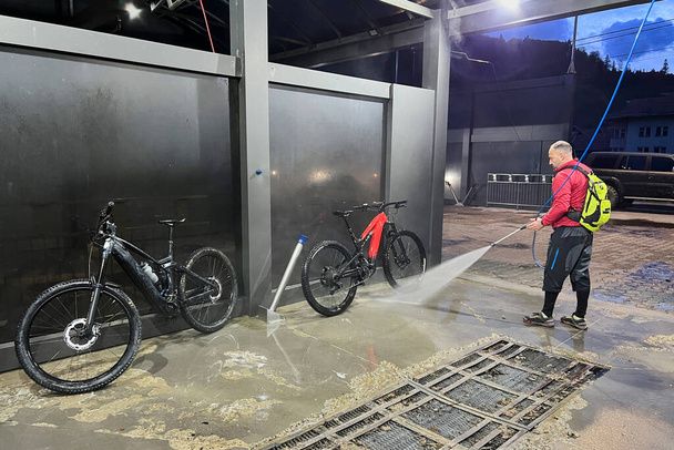 Ciclista usando spray de agua de alta presión para limpiar dos bicicletas eléctricas de montaña en la estación de lavado al aire libre. - Foto, imagen