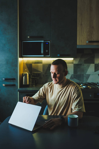 Молодой человек сидит за столом на кухне и работает на ноутбуке. Ноутбук на столе, а молодой человек работает дома.. - Фото, изображение
