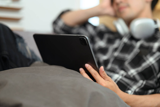 Unbekümmerter junger Mann liegt auf Couch und schaut sich zu Hause Filme auf digitalem Tablet an. - Foto, Bild