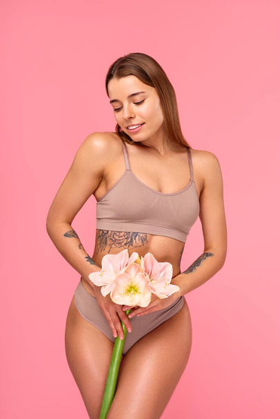 Perfect body. Slim caucasian lady posing with flower, showing her ideal curves, attractive woman posing in lingerie at pink studio background. Body and skin care, cosmetology concept. - Photo, Image