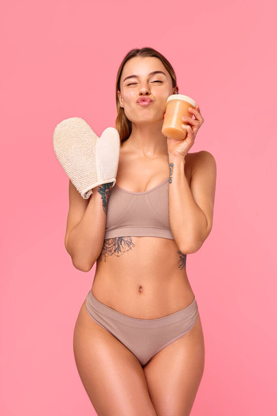 Karcsú, alsónadrágos nő masszírozza a test problémás területeit egy rózsaszín alapon izolált anti-cellulit kesztyűvel. Bőrápolás. Gyógykezelések és kozmetológia. - Fotó, kép