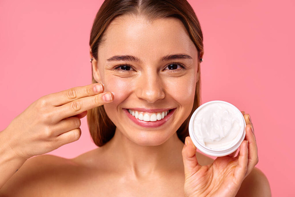 Caucasian woman with clear glowing skin using facial moisturizer or sunscreen isolated on pink background. Facial skin care. Spa treatments and cosmetology. - Photo, Image