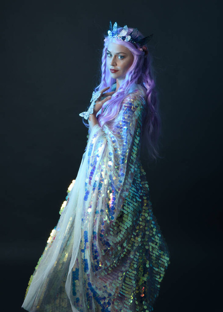 artistic portrait of beautiful female model with long purple hair wearing a fantasy fairy crown, wearing a rainbow glitter sequin ball gown. gestural flowing pose, isolated on dark studio background. - Photo, Image