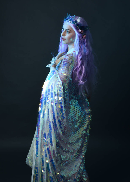 retrato artístico de la hermosa modelo femenina con el pelo largo de color púrpura con una corona de hadas de fantasía, con un vestido de lentejuelas arco iris brillo. pose gestual fluida, aislada sobre fondo oscuro del estudio. - Foto, imagen