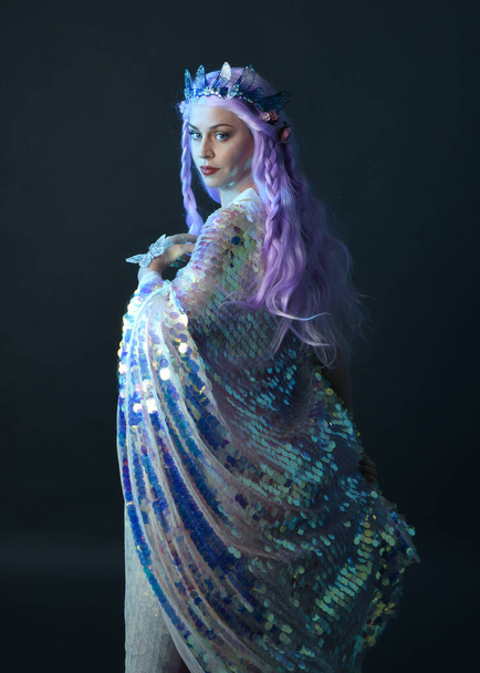 retrato artístico de la hermosa modelo femenina con el pelo largo de color púrpura con una corona de hadas de fantasía, con un vestido de lentejuelas arco iris brillo. pose gestual fluida, aislada sobre fondo oscuro del estudio. - Foto, imagen