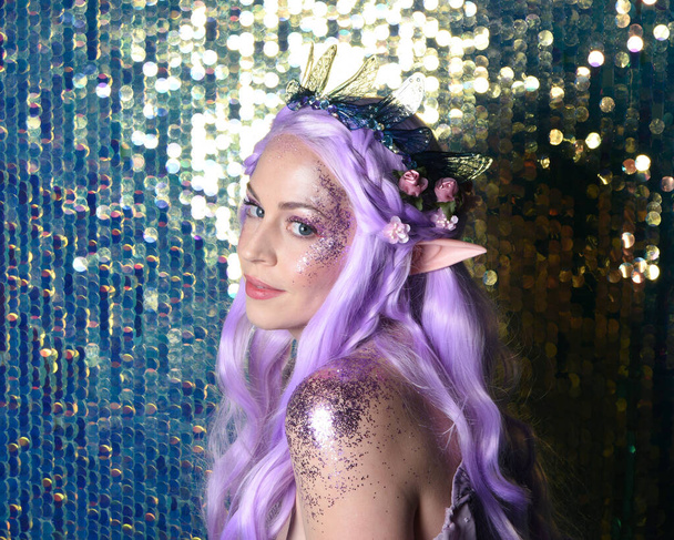  portrait of cute female model with long purple hair wearing a fantasy fairy flower crown with elf ears. Isolated on sparkling rainbow sequin background with glitter. - Photo, Image