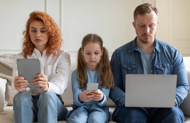 Problema de adicción a los gadgets. Los padres y la hija utilizando varios aparatos, ignorándose mutuamente, websurf con expresión aburrida en el sofá en casa - Foto, Imagen