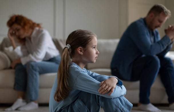 Unglückliches Mädchen, das unter Einsamkeit leidet, während ihre Eltern nach einem Streit nebeneinander auf dem Sofa sitzen, Beziehungsprobleme haben und sich selektiv auf ihre Tochter konzentrieren - Foto, Bild