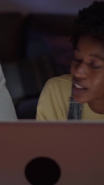 Verticaal. Jong multiraciaal koppel op zoek naar verkoop op de computer liggend op de vloer van de woonkamer 's nachts. Blanke man en Afrikaanse vrouw surfen online op laptop. Thuis leven van millennial tech mensen - Video