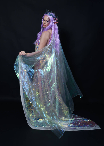 Full length πορτρέτο του όμορφου γυναικείου μοντέλου με μακριά μωβ μαλλιά φορώντας αυτιά ξωτικού, ένα μυθικό στέμμα και ουράνιο τόξο glitter πούλιες φόρεμα μπάλα. με τα πόδια, πίσω όψη, απομονωμένο σκοτεινό στούντιο  - Φωτογραφία, εικόνα