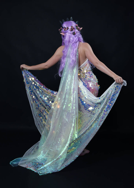 Full length πορτρέτο του όμορφου γυναικείου μοντέλου με μακριά μωβ μαλλιά φορώντας αυτιά ξωτικού, ένα μυθικό στέμμα και ουράνιο τόξο glitter πούλιες φόρεμα μπάλα. με τα πόδια, πίσω όψη, απομονωμένο σκοτεινό στούντιο  - Φωτογραφία, εικόνα