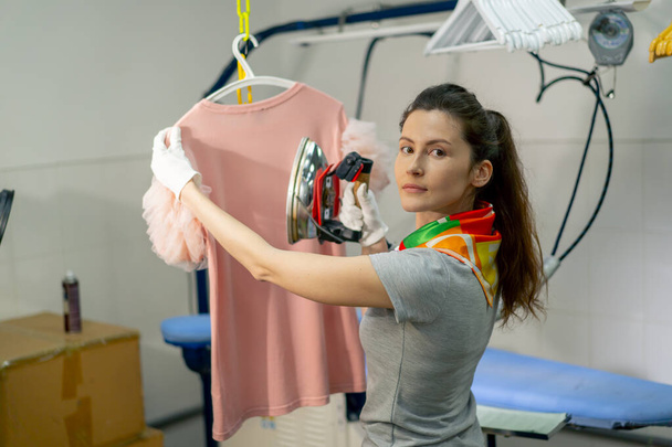 profesional de la limpieza en seco chica joven plancha un suéter rosa en una percha con mirada de vapor en la cámara - Foto, Imagen