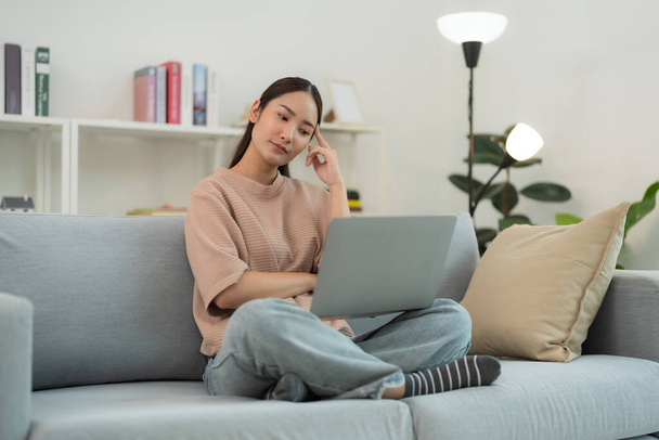 Młoda kobieta siedzi na szarej kanapie, patrząc skąpo na laptopa. Jest w swobodnym otoczeniu z relaksującą postawą, ucieleśnieniem zdalnej pracy lub badań online z przytulnym środowisku domowym - Zdjęcie, obraz