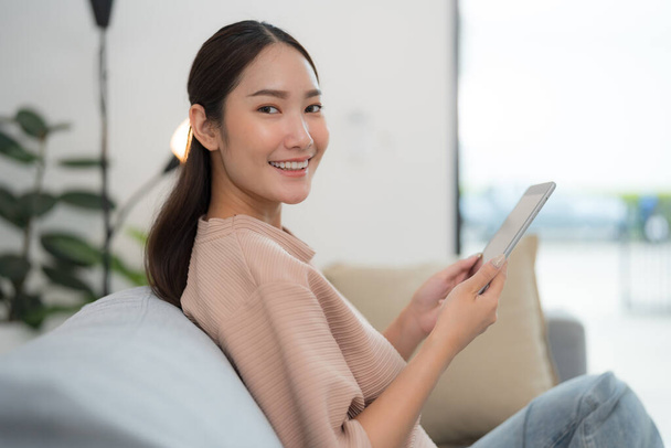 Schöne junge Asiatin sitzt gemütlich auf einer Couch und lächelt in die Kamera, während sie ein digitales Tablet in einem gut beleuchteten, modernen Wohnzimmer benutzt. - Foto, Bild