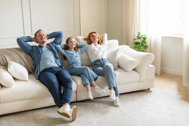 Relaxed parents and daughter resting on sofa with closed eyes and hands behind heads in living room interior, full length shot, free space - Photo, Image
