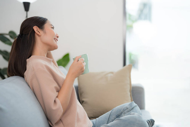 Młoda kobieta siedzi wygodnie na kanapie, trzymając kubek z delikatnym uśmiechem, odwracając wzrok z poczuciem zadowolenia w jasno oświetlonym nowoczesnym salonie - Zdjęcie, obraz