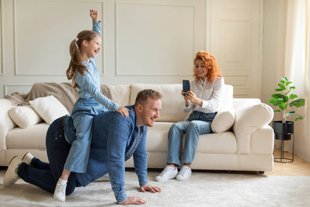 Family fun and games. Father playing with his daughter piggybacking girl, giving ride on his back while mother recording video on cellphone - Photo, Image