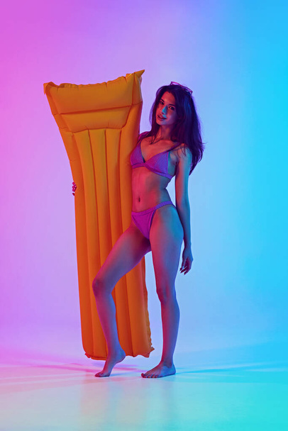 Nuori nainen täydellinen luku uimapuku poseeraa puhallettava neon valossa vastaan elinvoimainen kaltevuus tausta. Käsite kesälomat, matkustaminen, kauneus ja muoti, loma ja rentoutua. Ilmoitus - Valokuva, kuva