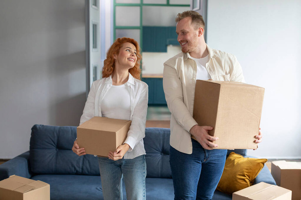 Концепция переселения. Позитивные супруги, несущие картонные коробки в собственном доме, переезжают в новую собственность - Фото, изображение