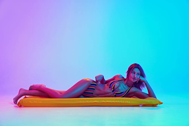 Young tender woman lying on air mattress and relaxing like swimming in ocean in neon light against vibrant gradient background. Concept of summer holidays, traveling, beauty and fashion, vacation. Ad - Photo, Image