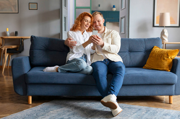 Rakastava eurooppalainen pari, joka jakaa sosiaalista mediaa kännykällä tai jolla on videopuhelu, onnelliset kumppanit sohvalla, mies syleilee naista - Valokuva, kuva