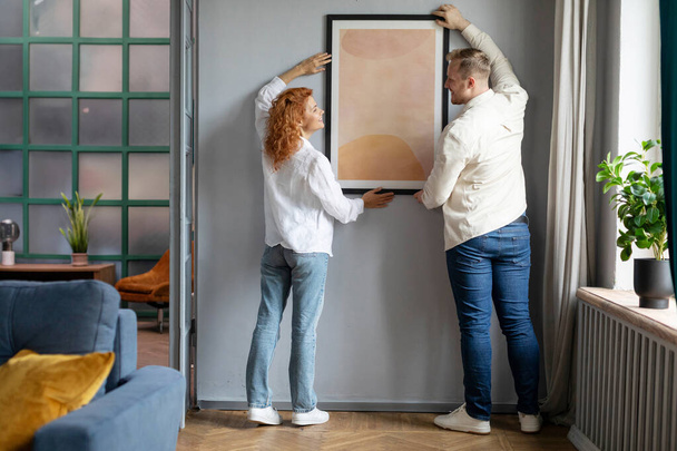 Conjoints accrochant l'affiche dans le cadre sur le mur, décorant leur intérieur de maison ensemble après la rénovation dans la maison neuve - Photo, image