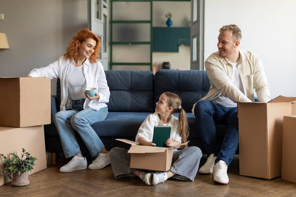 Cambio de casa. Familia europea de tres cajas de cartón desempaquetadas, sentadas en el interior de la sala de estar. Vivienda de madre, padre e hija - Foto, Imagen