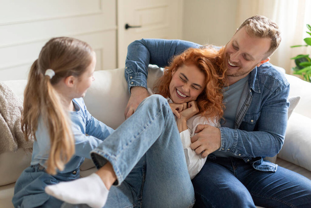 Радостная семья из трех человек веселится дома на диване, мужчина и девочка ухаживают за матерью и смеются, болтая по интерьеру гостиной - Фото, изображение