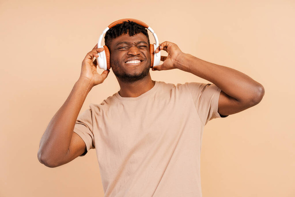 Aantrekkelijke Afrikaanse jonge hipster man met draadloze hoofdtelefoon luisteren naar muziek geïsoleerd op beige achtergrond op zoek naar camera genieten van muziek, moderne technologie concept - Foto, afbeelding