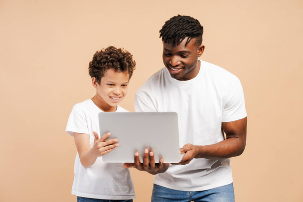 Izgalmas, boldog afro-amerikai férfi, apa és az ő kis aranyos fia, laptoppal, rendelés, online vásárlás, videó nézése, játék, álló elszigetelt bézs háttér - Fotó, kép