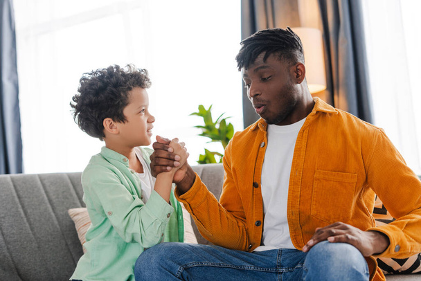 Happy, handsome, African American father and little son playing, checking who is stronger, sitting on comfortable sofa at home. Concept of childhood, parenthood - Photo, Image
