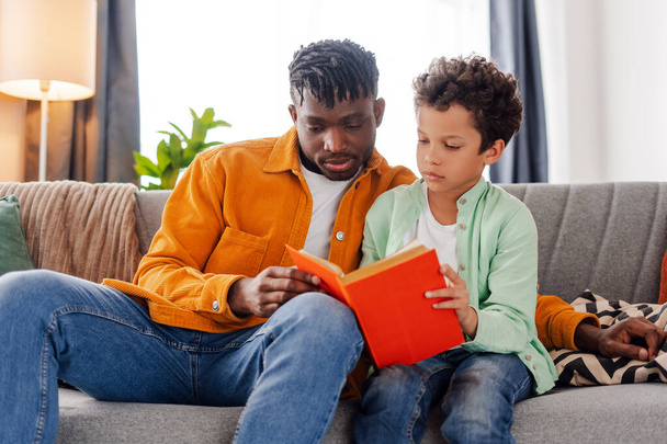 Nuori, huolehtiva afroamerikkalainen isä lukee kirjaa pienen poikansa kanssa istuessaan mukavalla sohvalla kodikkaassa kodissa. Opiskelun, koulutuksen, harrastuksen käsite - Valokuva, kuva