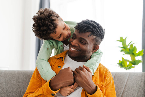 Smiling, attractive African American young father and little son hugging, sitting on comfortable sofa in cozy home. Concept of parenthood, relationships - Photo, Image