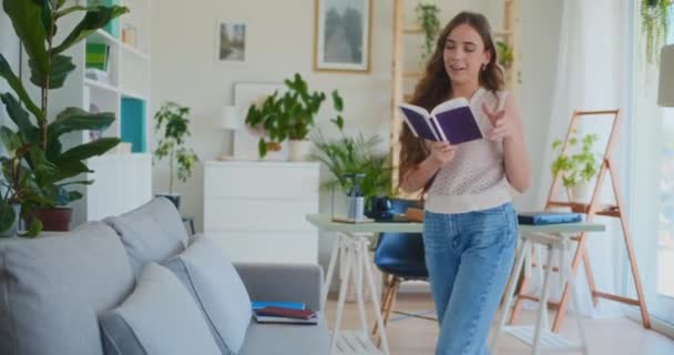 Χαρούμενη γυναίκα διαβάζει ενώ περπατάει και κάθεται στον καναπέ της στο σπίτι - Πλάνα, βίντεο