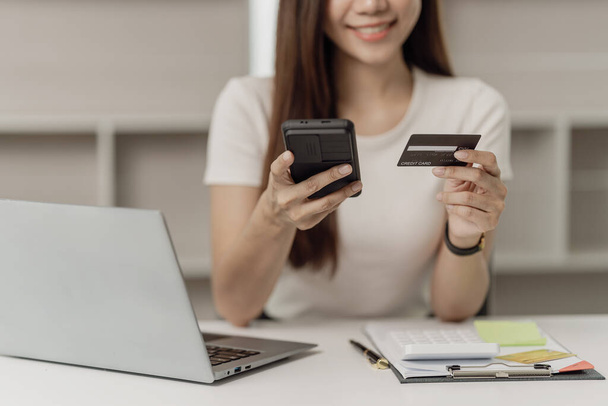 Atractiva mujer asiática sostiene teléfono inteligente y tarjeta de crédito en su computadora portátil utilizando la banca móvil o la aplicación de compras en líneaPago a través de la banca por Internet - Foto, imagen