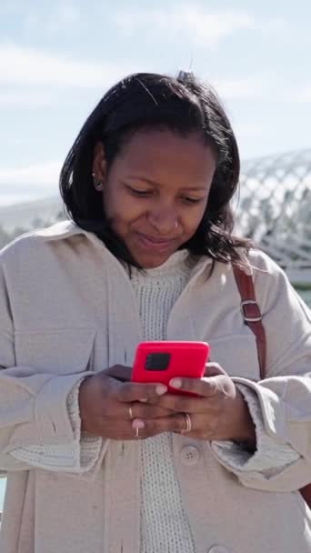 Vertical. mujer latina sonriente utilizando y mirando teléfono móvil inclinada barandilla en el parque científico de la ciudad de Valencia. Turista negra escribiendo en la celda en el soleado día de invierno. Comunicarse en viajes - Metraje, vídeo