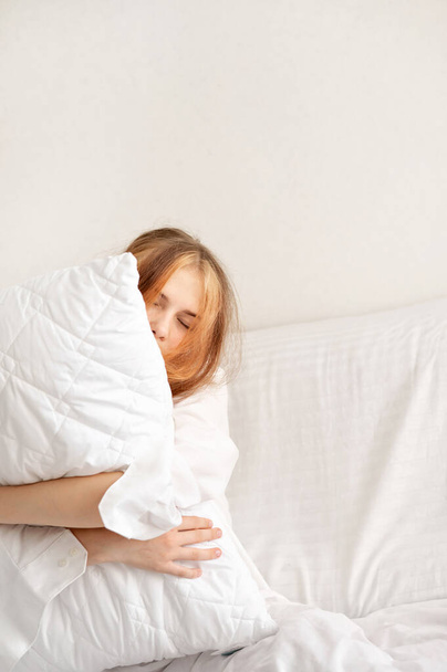 Sleepy teenage girl with eyes closed embracing white pillow in neutral white bedroom, teenagers sleep deficiency, good sleep concept. - Photo, Image