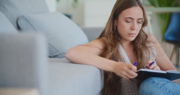 Woman starts to write in a notebook while sitting by the sofa at home learning - Footage, Video