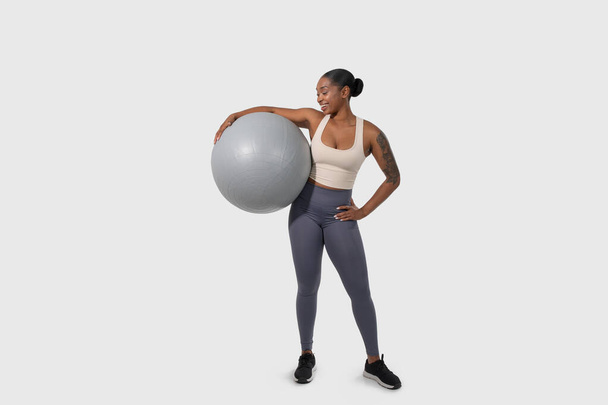 La mujer afroamericana sostiene una pelota de ejercicio frente a un fondo blanco. Ella está de pie con confianza, agarrando la pelota con ambas manos, con una expresión enfocada. - Foto, imagen