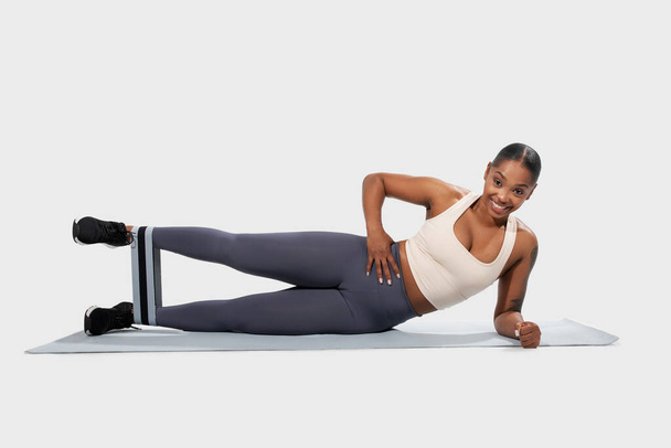 Веселая афро-американка в спортивной одежде держит боковую доску позы с бандажом сопротивления на ногах, привлекая свои основные мышцы - Фото, изображение