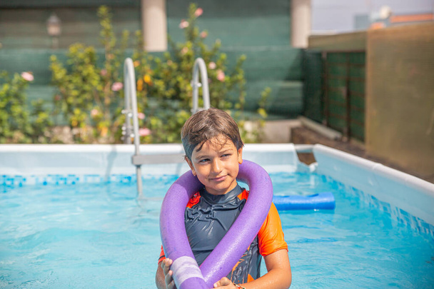 Веселый мальчик в оранжевой рубашке с фиолетовой плавательной лапшой в ярком солнечном открытом бассейне - Фото, изображение