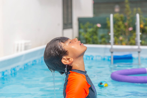 Chłopiec w pomarańczowej koszuli pływackiej lubi się ochłodzić, głowa odchylona z powrotem z kaskadą wodną, makaronem i schodami basenowymi - Zdjęcie, obraz