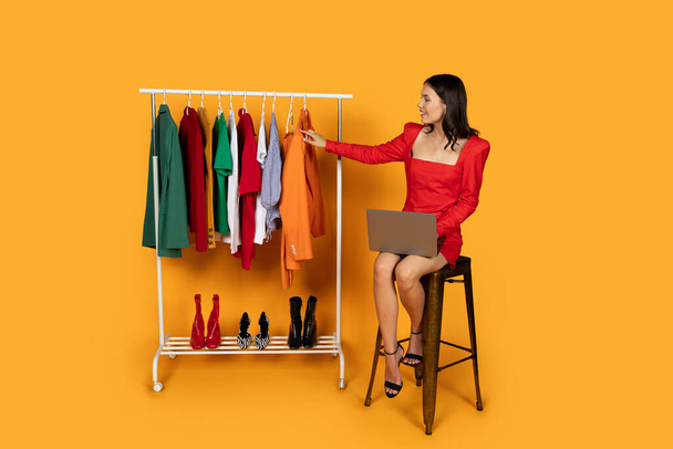 A stylish woman sits on a tall stool, holding a laptop, while thoughtfully examining a selection of colorful autumn clothing hung on a rack, with a range of shoes lined up below - Photo, Image