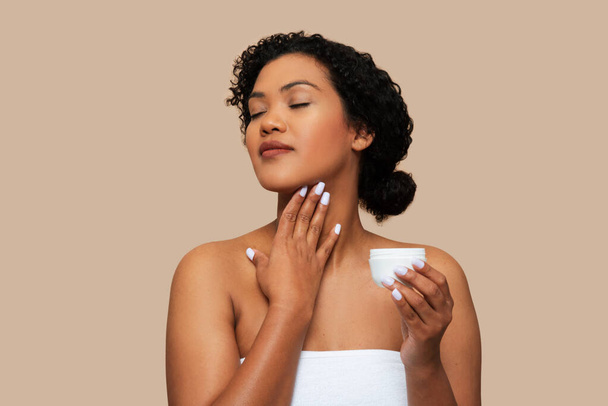 A young woman with curly hair, wrapped in a white towel, gently applies moisturizing cream to her neck, moment of self-care and relaxation. The neutral beige background - Photo, Image
