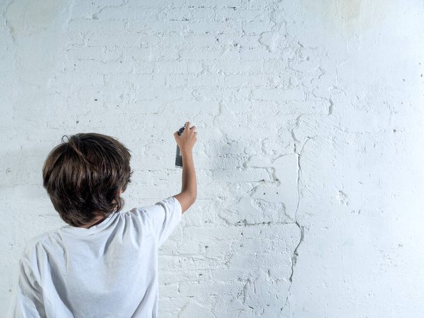 Poika maalaa seinää suihkepullolla. Pojalla on valkoinen paita ja ruskeat hiukset. Poika on keskittynyt tehtäväänsä ja näyttää nauttivan - Valokuva, kuva