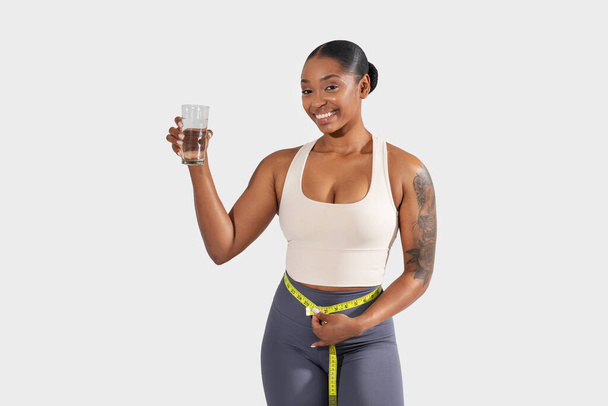 Μια χαρούμενη μαύρη γυναίκα στέκεται με αυτοπεποίθηση με ένα μέτρο ταινία γύρω από τη μέση της, κρατώντας ένα καθαρό ποτήρι νερό. Το ενεργητικό της ένδυμα υποδεικνύει μια εστίαση στην υγεία και την καλή φυσική κατάσταση - Φωτογραφία, εικόνα
